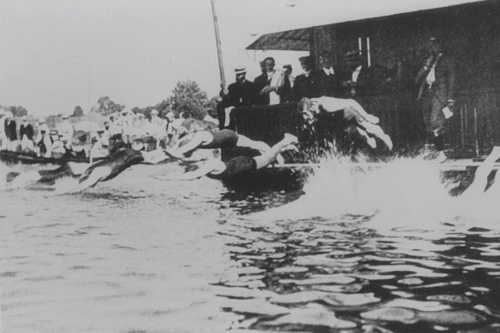Historical black-and-white photo of multiple swimmers diving into a body of water, marking the beginning of a swimming race. Onlookers dressed in vintage clothing and hats are gathered on the dock, observing the competition
